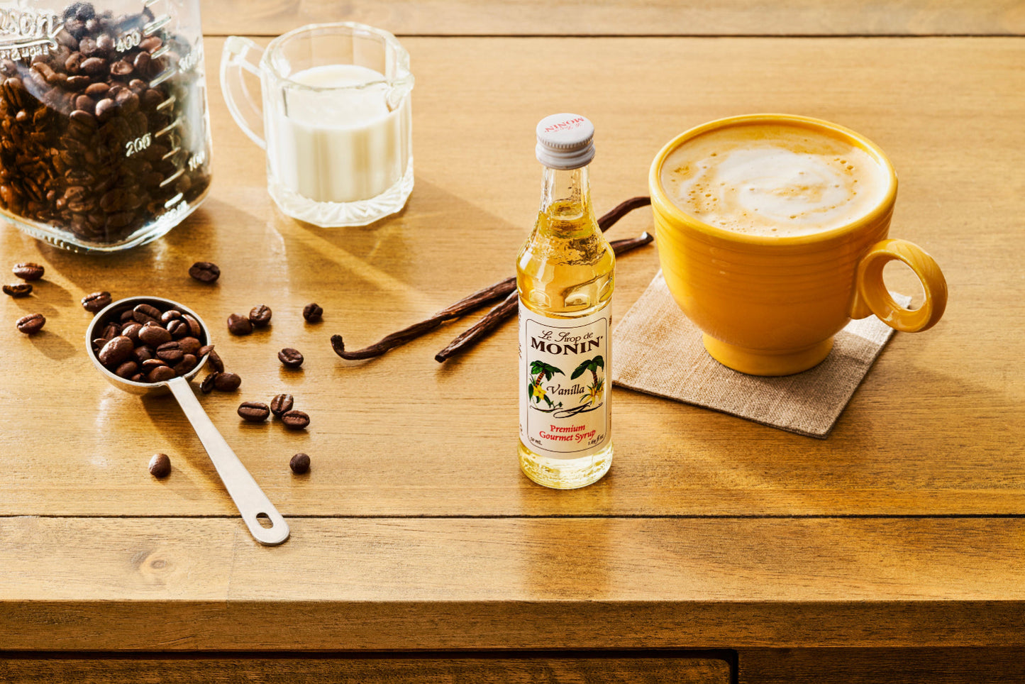 A sample-size 50 mL Monin Vanilla Syrup bottle sits beside a yellow mug filled with a frothy latte. A tablespoon filled with coffee beans, vanilla bean pods, and a pitcher of milk sit on the wooden table.