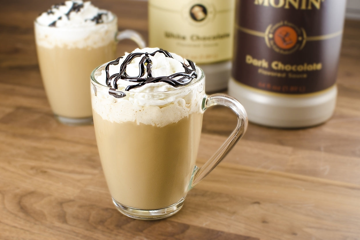 A cup of hot chocolate with bottles of white chocolate and dark chocolate Monin syrup in the back