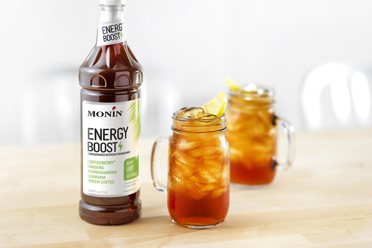 Monin Energy Boost bottle next to two iced beverages in mason jars garnished with lemon slices on a wooden surface.