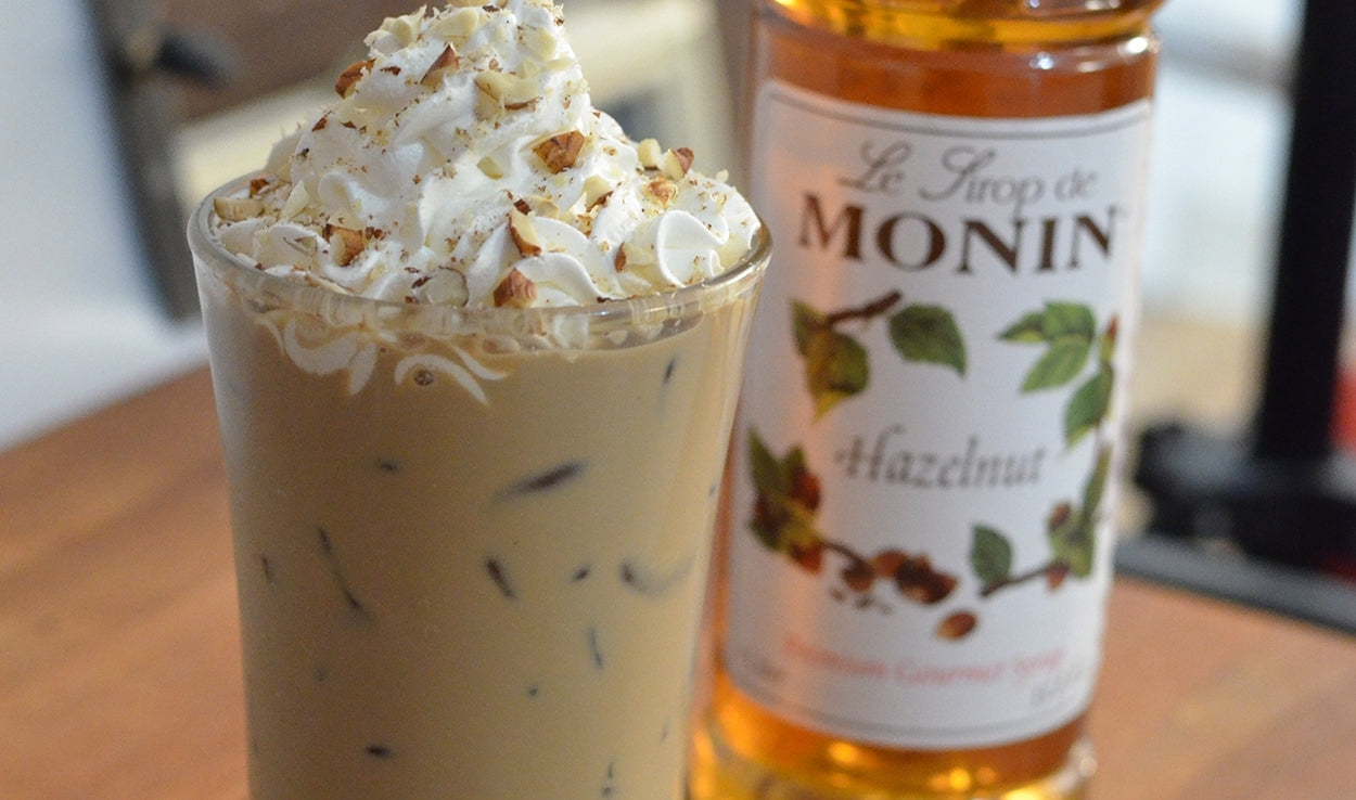 close up of iced drink with whipped cream using Monin hazelnut syrup with the bottle of Hazelnut in the background