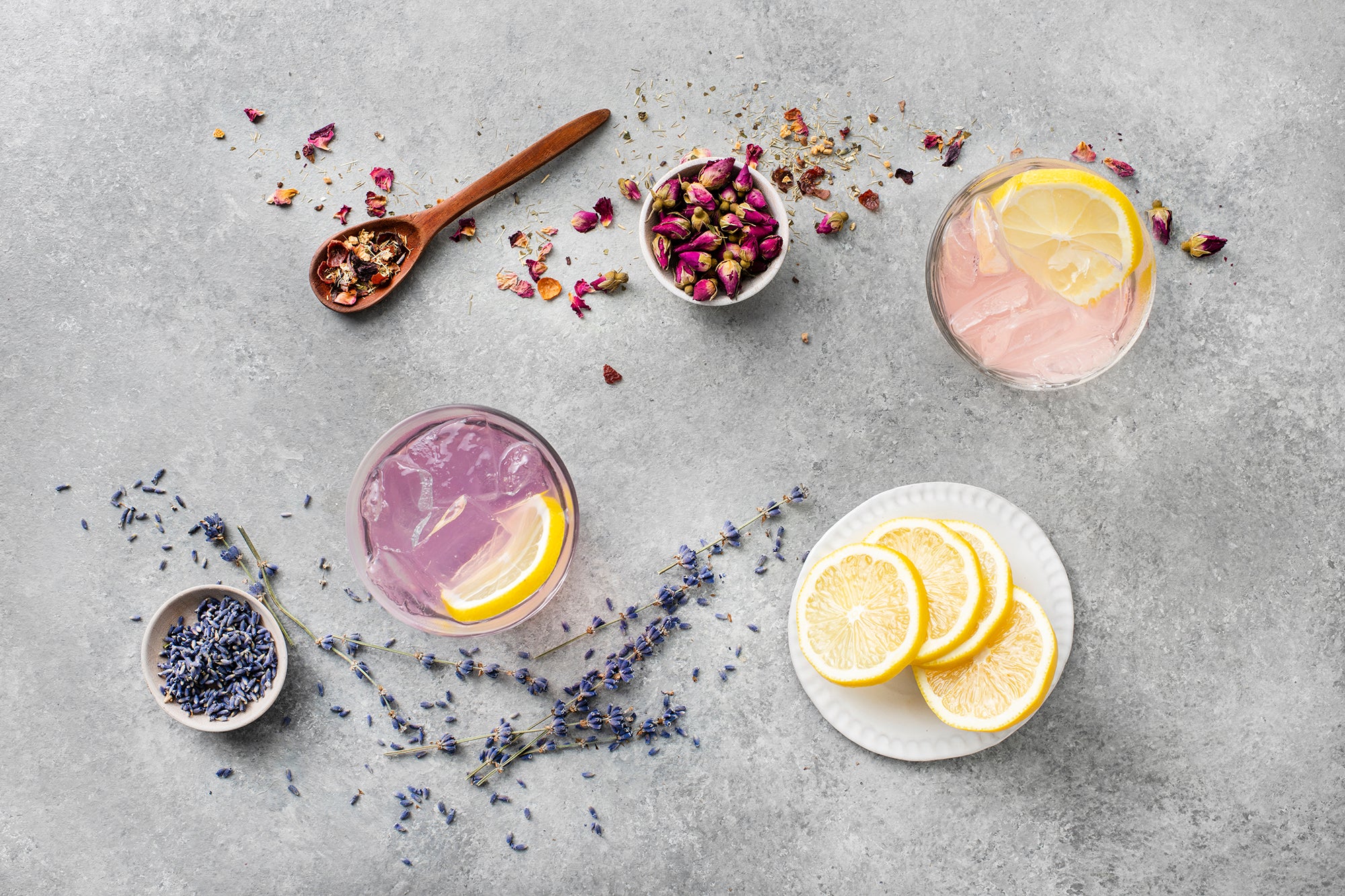 Overhead shot of purple and pink iced drinks with lavender and rose petals along with a plate of lemons.