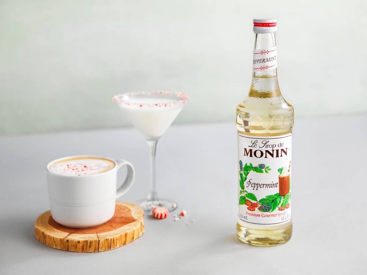 A hot latte and a peppermint martini sitting next to a bottle of Monin Peppermint Syrup.