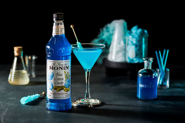 A blue Cotton Candy Martini sitting in front of a chemistry set up filled with blue liquid. All on a black backdrop. Easy Halloween drink to make for a party.