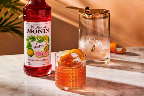 In the forefront is a rocks glass filled with orange-pink liquid and garnished with an orange peel. In the background is a bottle of Guava Syrup and an empty mixing glass with melted ice in it