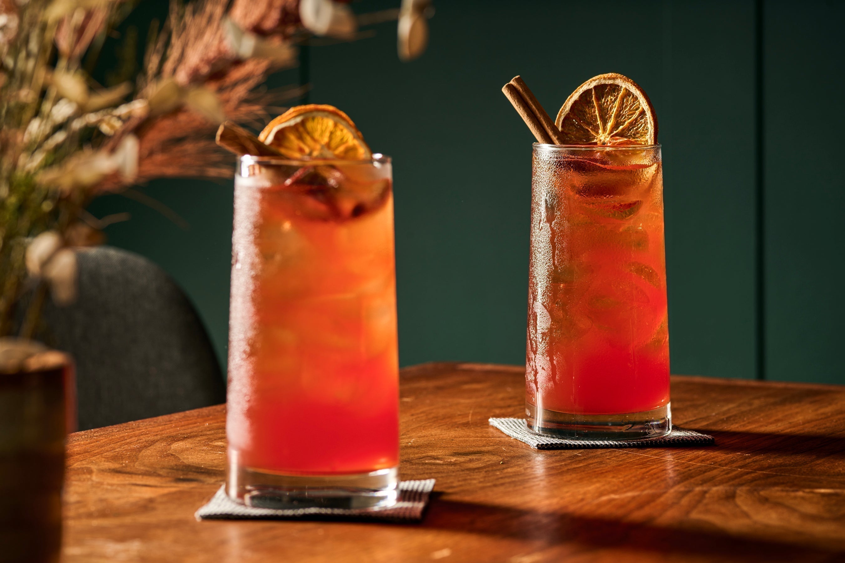 Two tall glasses filled with bright orange liquid, they are easy Halloween drinks made with Monin Pumpkin Spice Syrup and garnished with cinnamon stick and dried orange wheels.