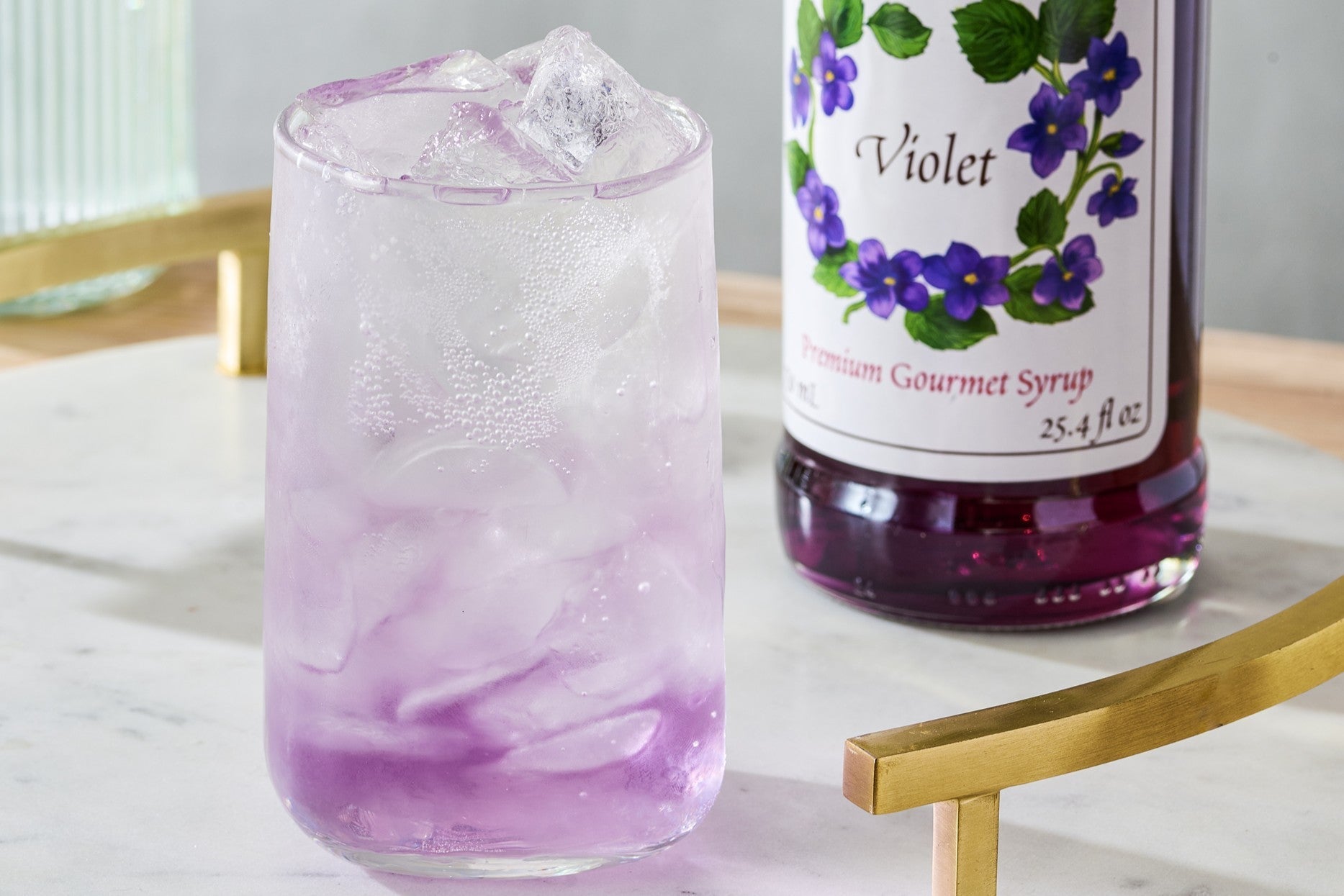 A violet spring cocktail sitting on a marble tray, with a bottle of Monin Violet Syrup in the backgroud.