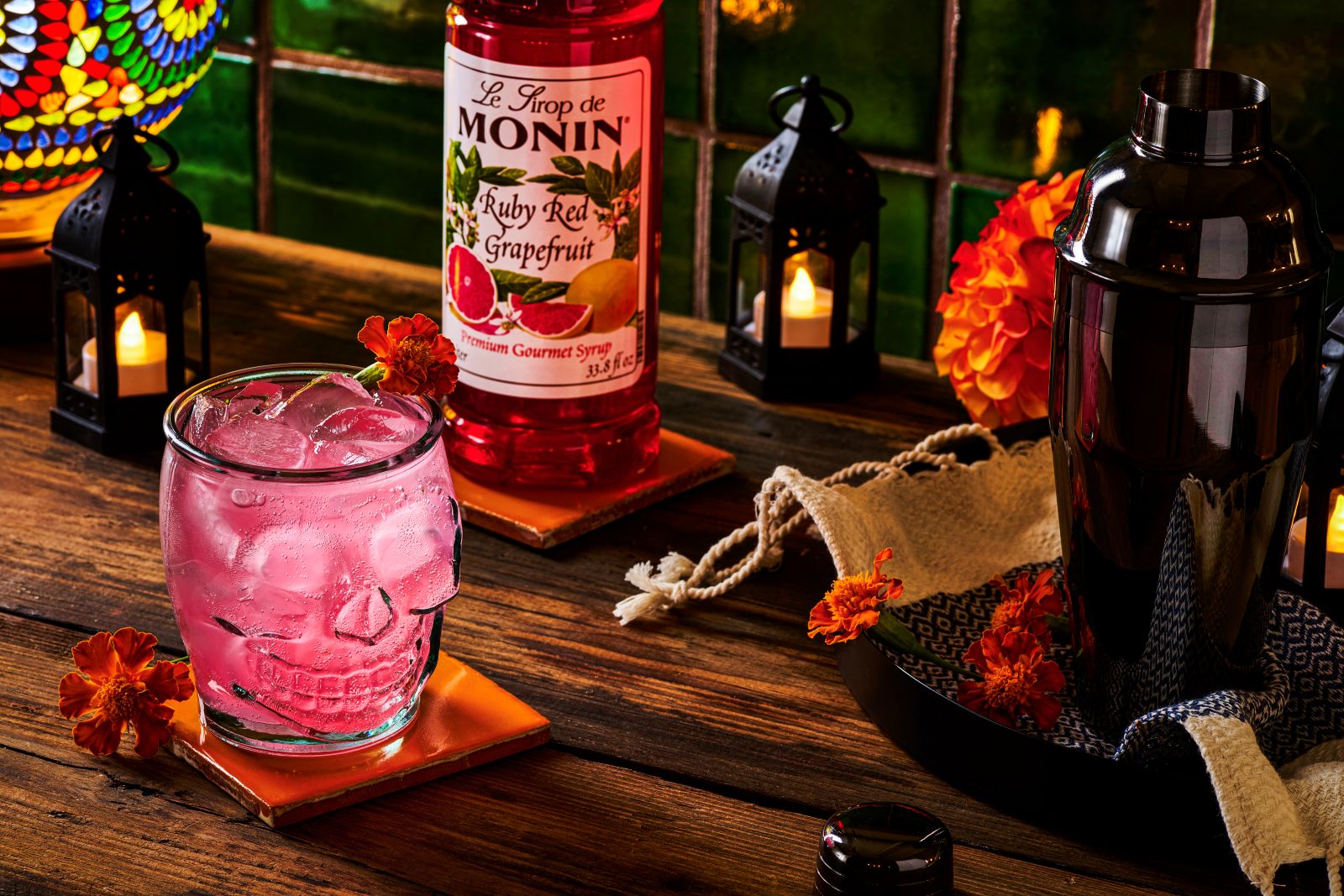 Pink grapefruit cocktail served in a glass skull, surrounded by marigolds, candles, and Monin Ruby Red Grapefruit Syrup — Day of the Dead–inspired drink setup.
