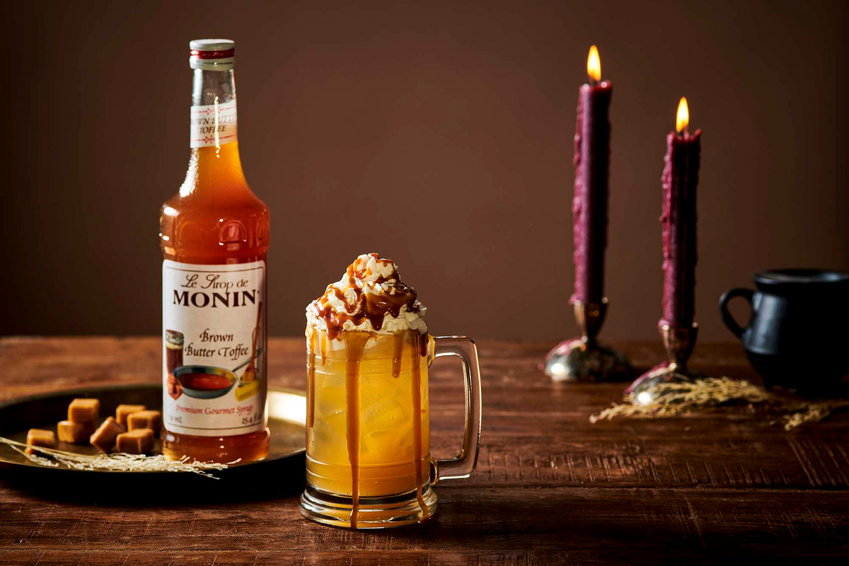 Iced caramel drink topped with whipped cream and toffee drizzle in a glass mug, alongside a bottle of Monin Brown Butter Toffee Syrup and lit candles.