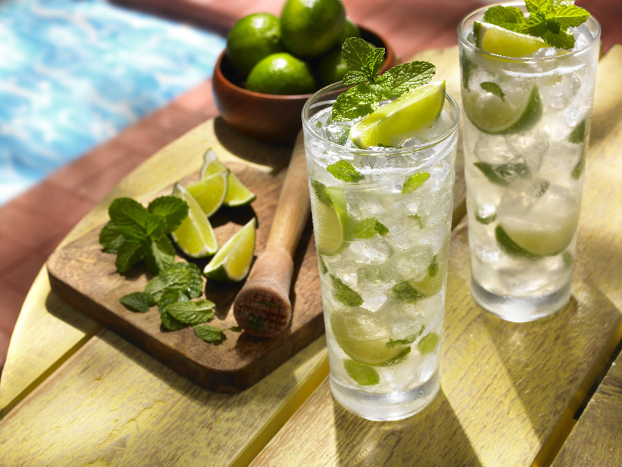 Two poolside cocktails (mojitos) next to a muddler, lime wedges, and mint leaves.