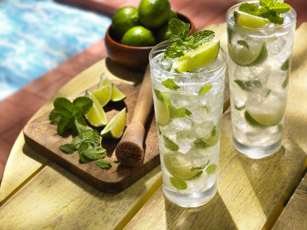 Two poolside cocktails (mojitos) next to a muddler, lime wedges, and mint leaves.