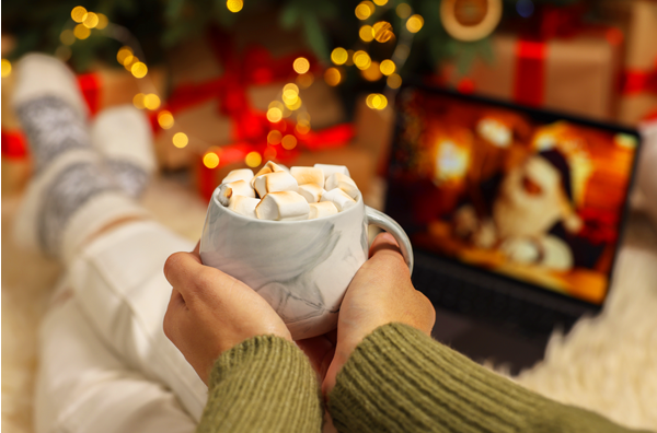 Two hands holding a mug of hot chocolate with toasted marshmallow on top, sitting in front of a laptop and watching a holiday movie.