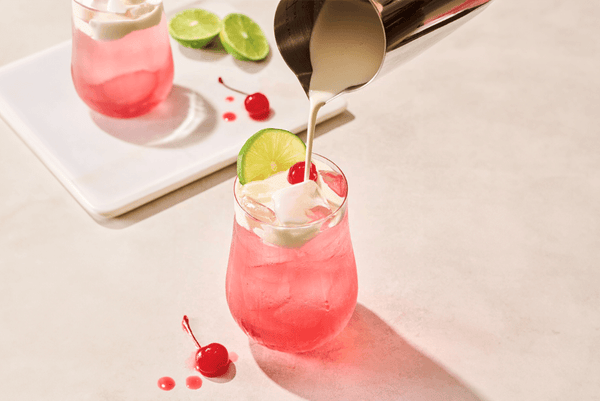 In the foreground, a pink soda garnished with a lime wheel is being topped with cream from a pourer. Another pink dirty soda, cherries, and limes are on a platter in the background.