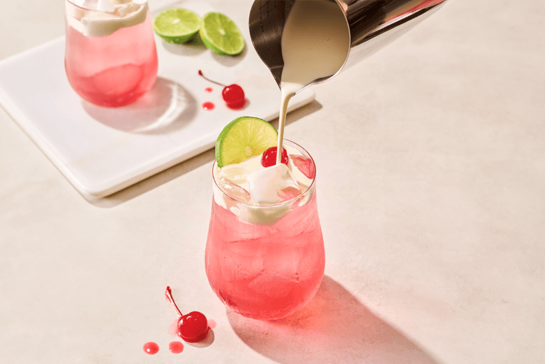 In the foreground, a pink soda garnished with a lime wheel is being topped with cream from a pourer. Another pink dirty soda, cherries, and limes are on a platter in the background.