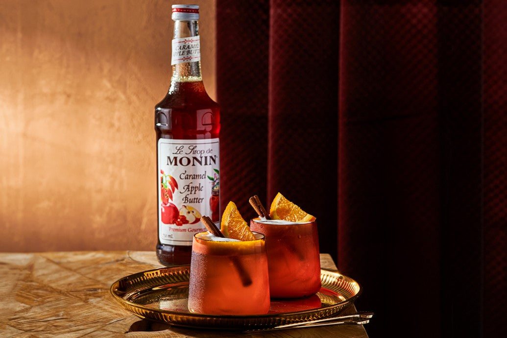 A bottle of Monin Caramel Apple Butter Syrup displayed beside two cocktails on a gold tray. The drinks are garnished with orange slices and cinnamon sticks, highlighting a warm, autumn-inspired presentation.