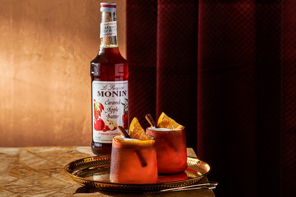 A bottle of Monin Caramel Apple Butter Syrup displayed beside two cocktails on a gold tray. The drinks are garnished with orange slices and cinnamon sticks, highlighting a warm, autumn-inspired presentation.
