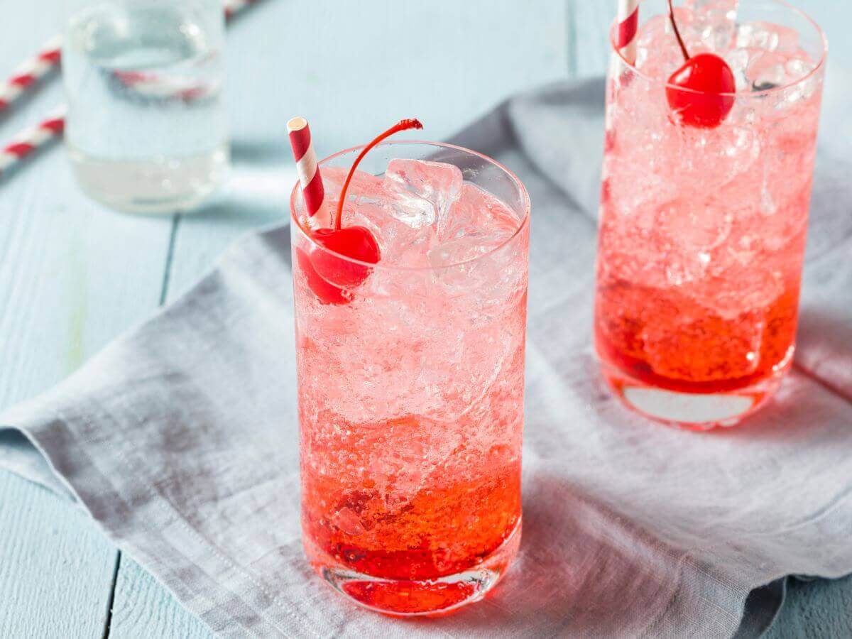 two glasses filled with pink liquid, ice, and garnished with cherries. They are sitting on a gray napkin placed upon a wooden table.