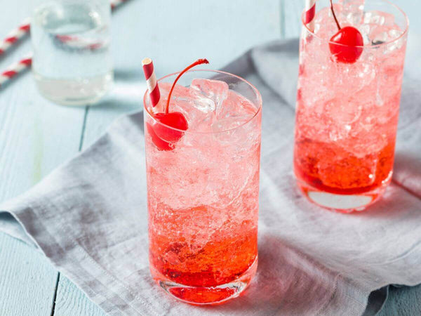 two glasses filled with pink liquid, ice, and garnished with cherries. They are sitting on a gray napkin placed upon a wooden table.