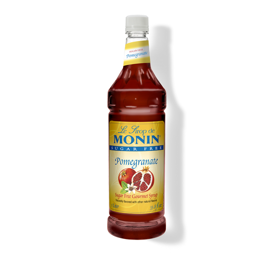 A bottle of Monin sugar-free pomegranate syrup on a white background with a soft shadow. The bottle is filled with deep red syrup and features a yellow label with blue and red text, along with an illustration of a pomegranate.
