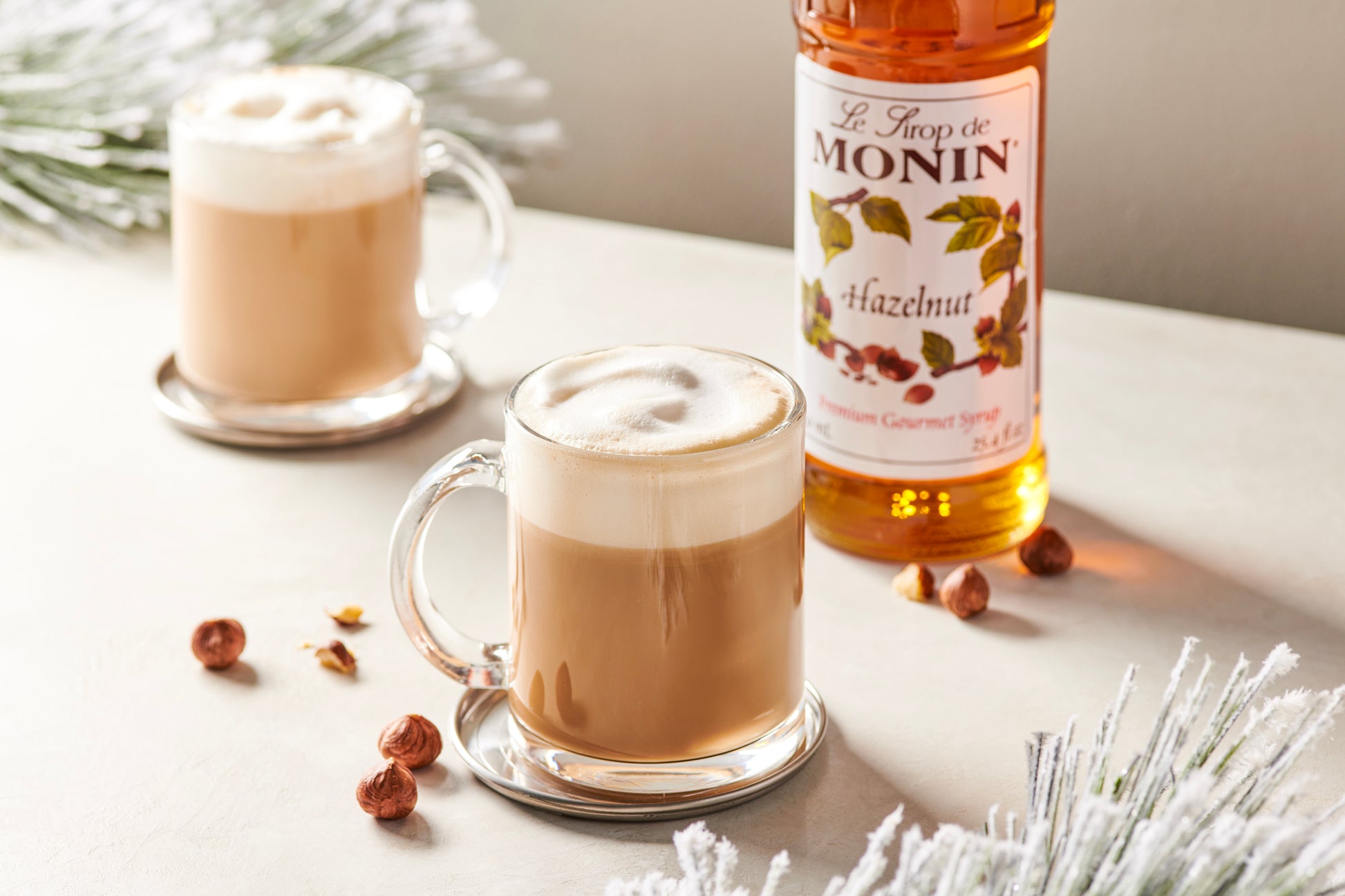 Two glasses of hazelnut latte with a bottle of Monin hazelnut syrup on a white surface.