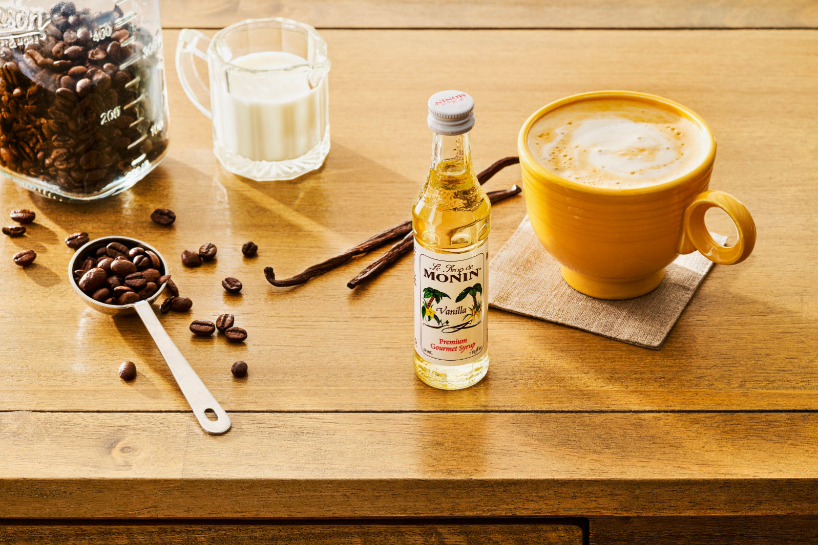 A sample-size 50 mL Monin Vanilla Syrup bottle sits beside a yellow mug filled with a frothy latte. A tablespoon filled with coffee beans, vanilla bean pods, and a pitcher of milk sit on the wooden table.