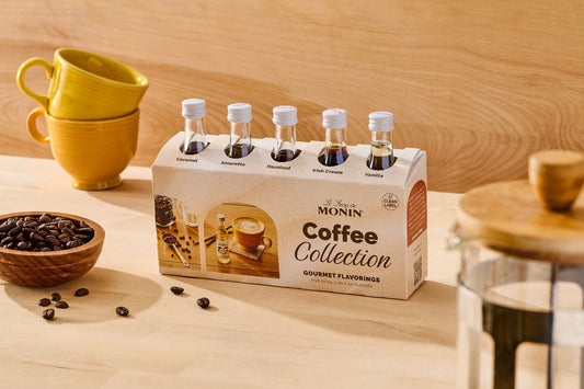 A Monin Coffee Collection gourmet flavorings gift set with 5 mini bottles of Caramel, Amaretto, Hazelnut, Irish Cream and Vanilla Syrups is displayed on a wooden table with coffee beans in the foreground, and yellow coffee cups in the background.