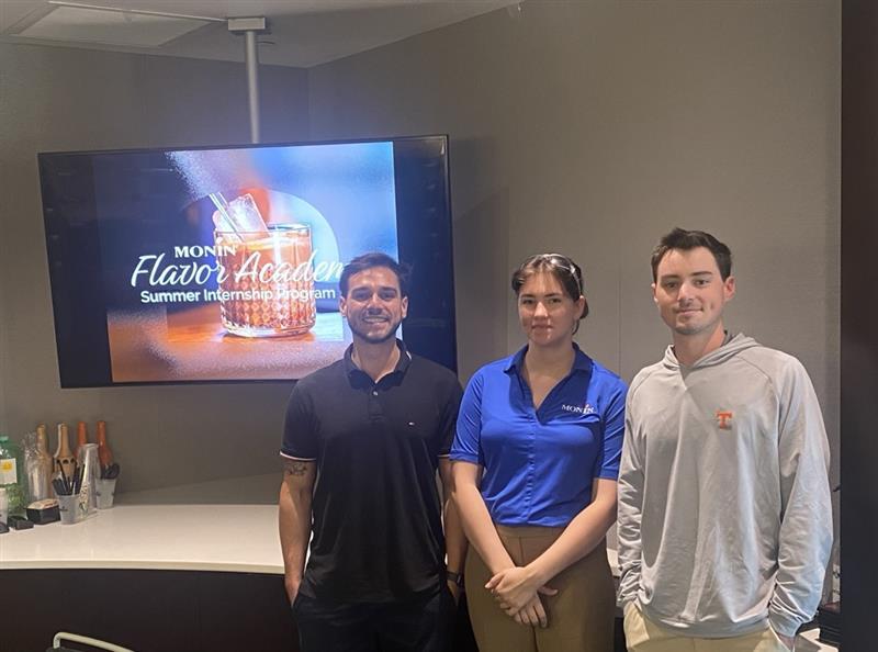 Three people standing in front of a screen displaying a Monin Flavors Academy advertisement.