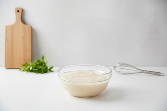 Clear glass bowl with creamy white sauce on a light gray surface, with a wooden cutting board and whisk in the background.