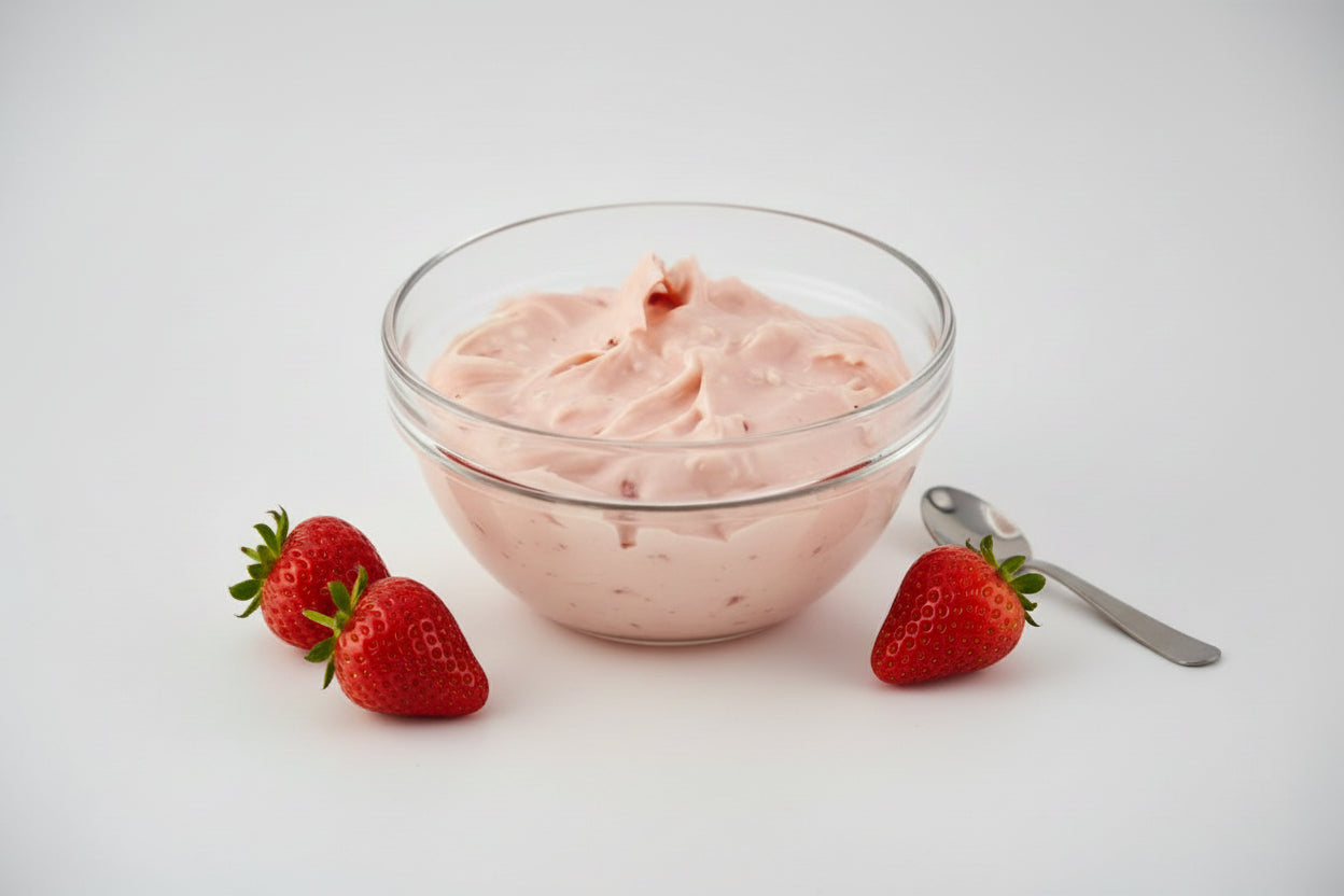 Pink cream in a glass bowl on a white background with a small spoon and a few strawberries on the side. 