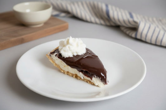 Slice of peanut butter pie, topped with chocolate and with whipped cream on a white plate, placed on a gray surface with a wooden cutting board and striped cloth in the background.