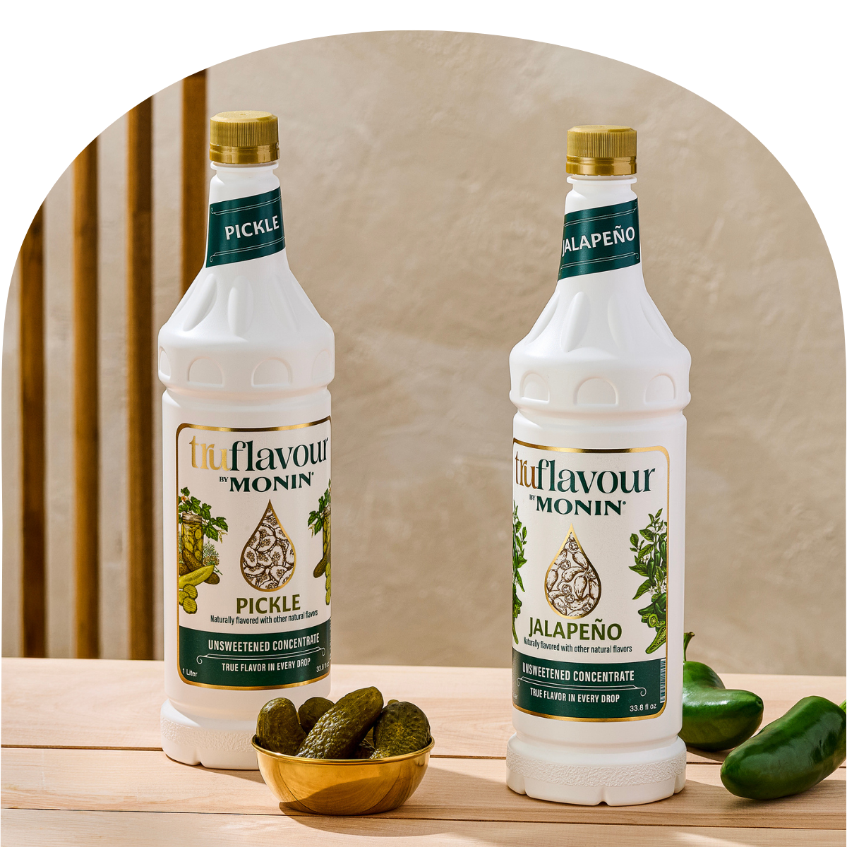 Two bottles of Monin Flavour Concentrate on a wooden surface with pickles and jalapeño peppers.