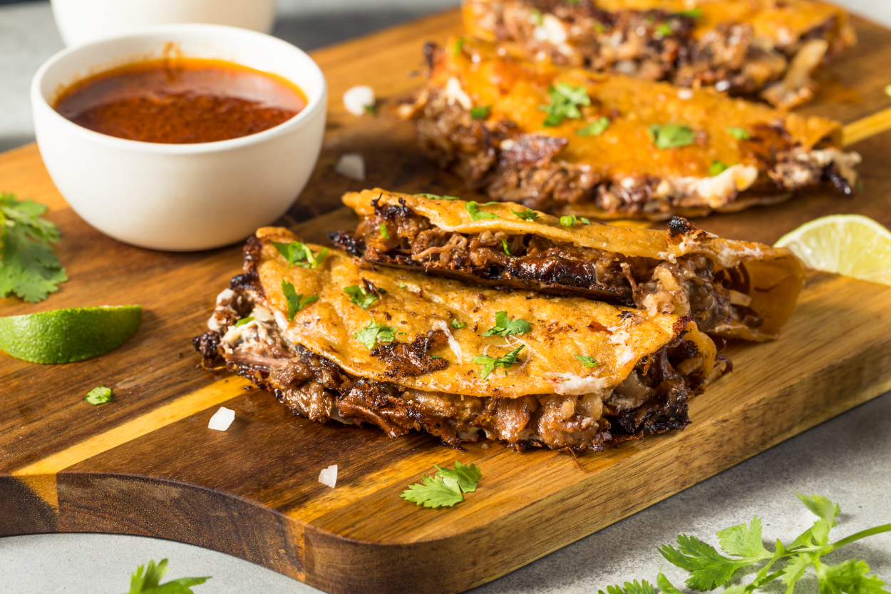 a cutting board with four tacos on it next to some birria sauce