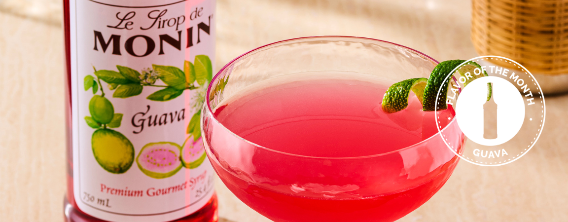 Monin Guava syrup bottle with a glass of pink guava juice on a wooden surface.