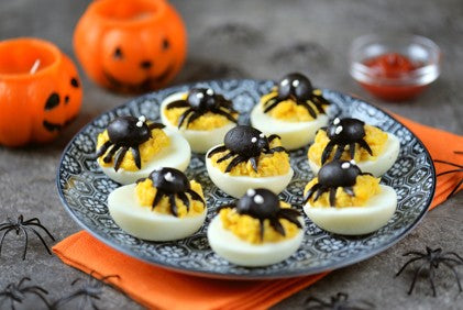 Deviled eggs decorated with spiders on a Halloween-themed plate with pumpkins and spider decorations around