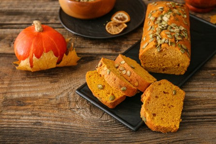 Loaf of pumpkin bread with slices on a dark plate, surrounded by pumpkins and dried oranges on a wooden surface.